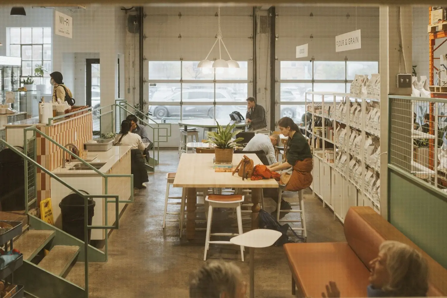 People working and talking at a long shared table in the Almanac café, under tall industrial windows.